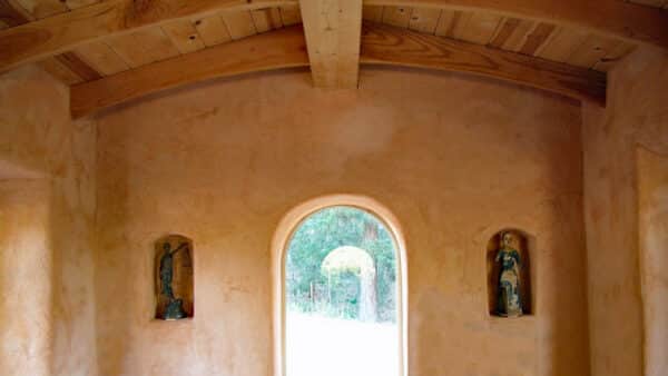 Strawbale Cottage - Sunset Cottage interior looking out an arched window set in a natural plaster wall with two niches and wood ceiling.