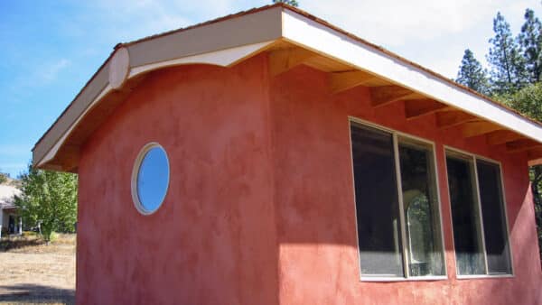 Strawbale cottage - Sunset Cottage almost completed. View of round window and window seat from exterior with red lime plaster finish coat.